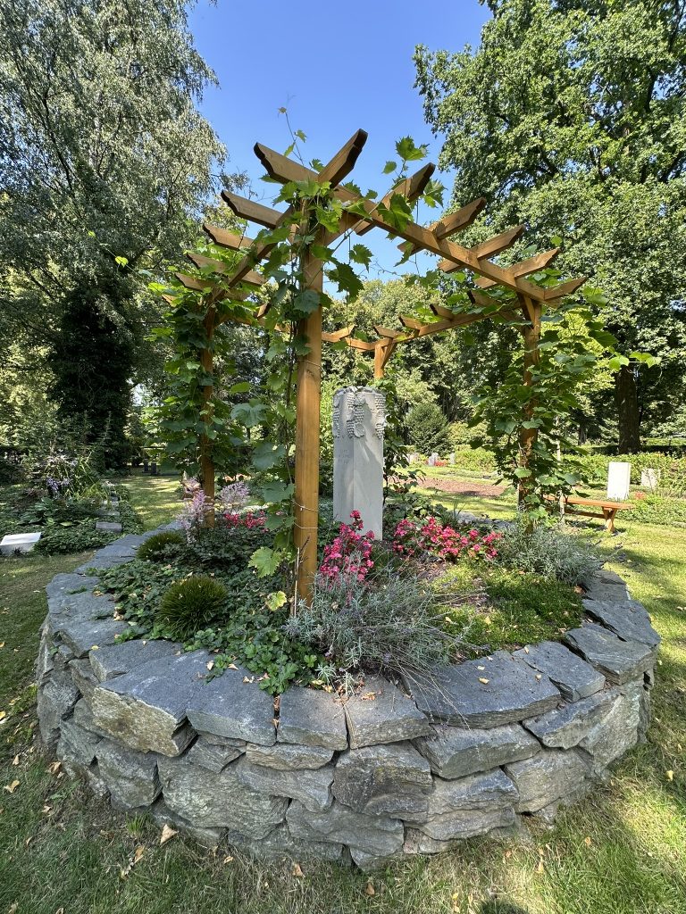 Rondell mit Weinpergola (Herzstück). Der echte Wein steht für Leben, Zusammenhalt und Erinnerung. Natursteinmauer und Namensstein bilden einen würdigen Rahmen.