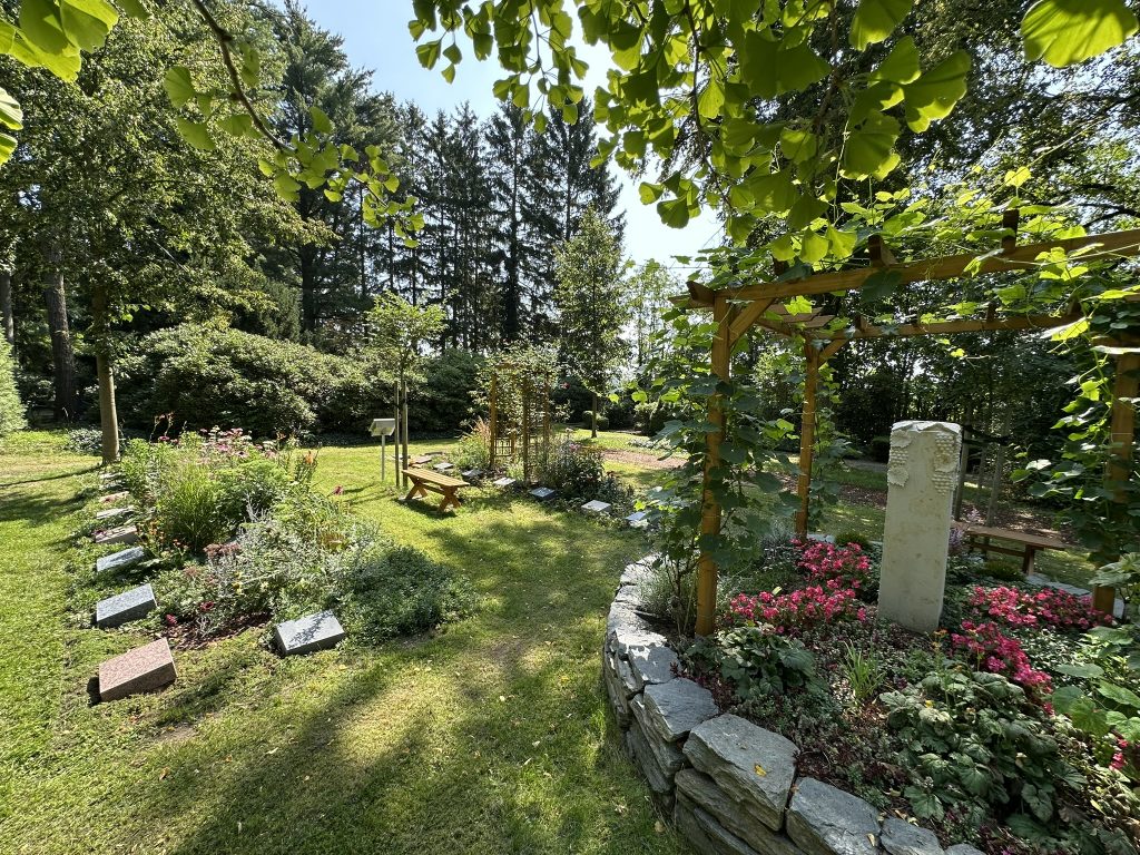 Zentrales Rondell mit Natursteinmauer und Weinpergola; Namensstein als Ort gemeinsamer Erinnerung.